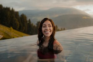 A young woman smiles while looking off camera. She's standing in an infinity poll outside with mountains, trees, and sky behind her, in this Lemonaid Health article answering the question, "are compounded GLP-1s safe?"